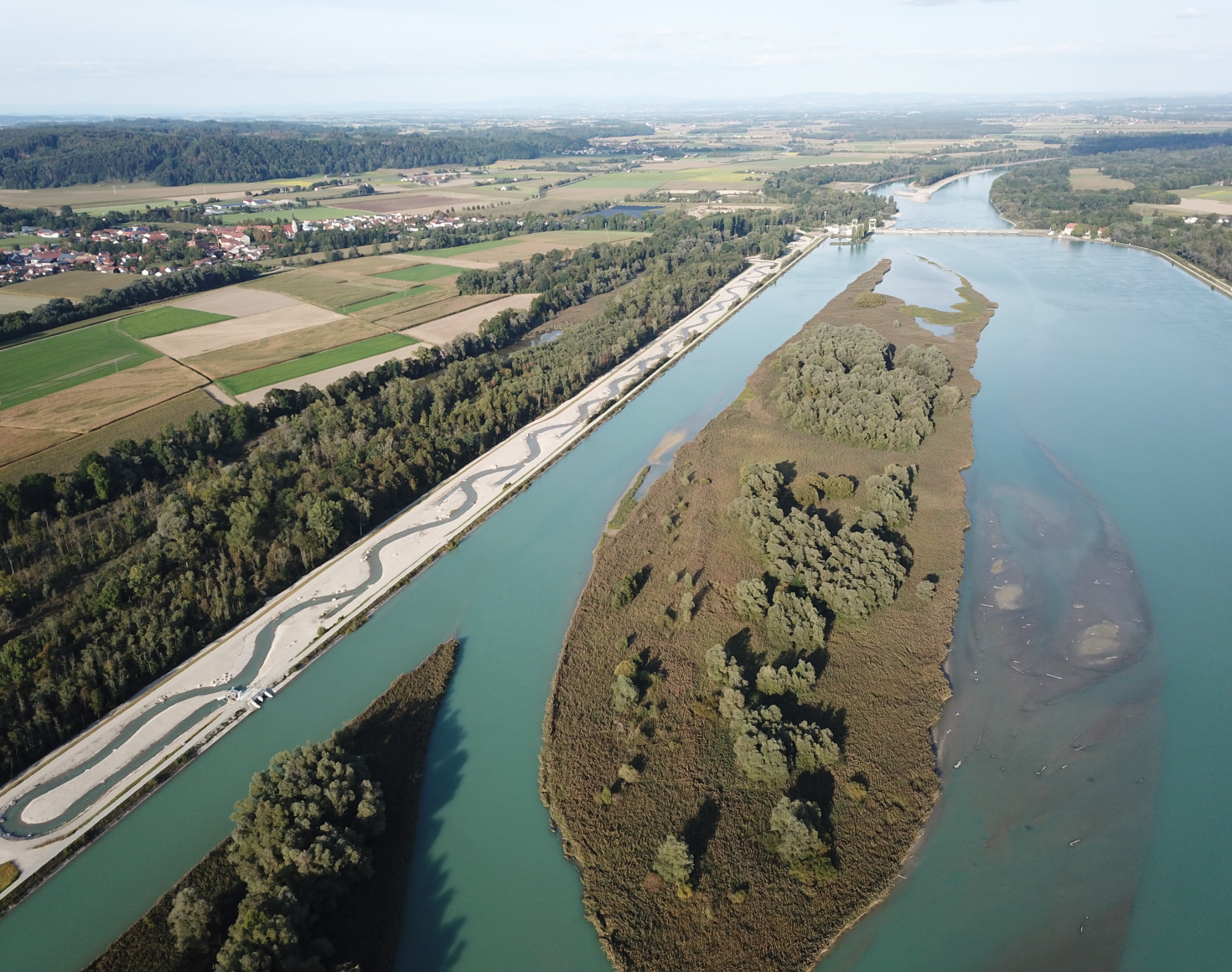 The aerial photo shows the section of the Inn before the Ering-Frauenstein Inn power station. The fish migration aid can be seen on the left-hand side of the image next to a section of forest. In the centre of the image is the renaturalised section of the Inn, which shines in a dark blue. The renaturalised area is fully overgrown with trees and bushes. In the background of the picture you can see the further course of the Inn as well as the landscape with villages and fields in various colours.