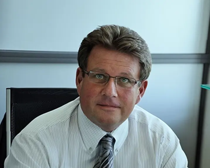 Roland Schmalfuß poses in his office. He is wearing glasses, a striped shirt and a tie.